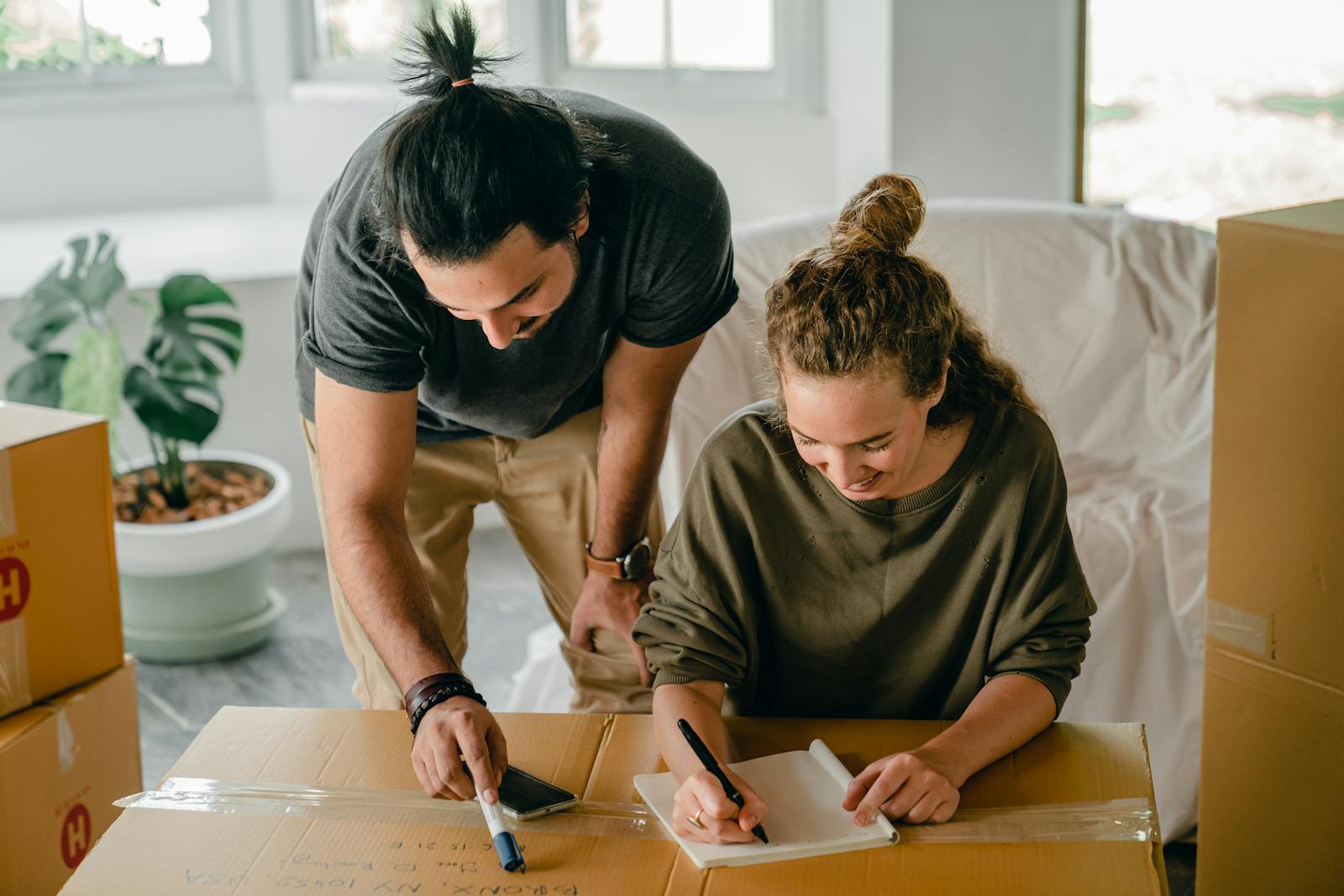 Cheerful diverse couple writing in notebook near boxes before relocation